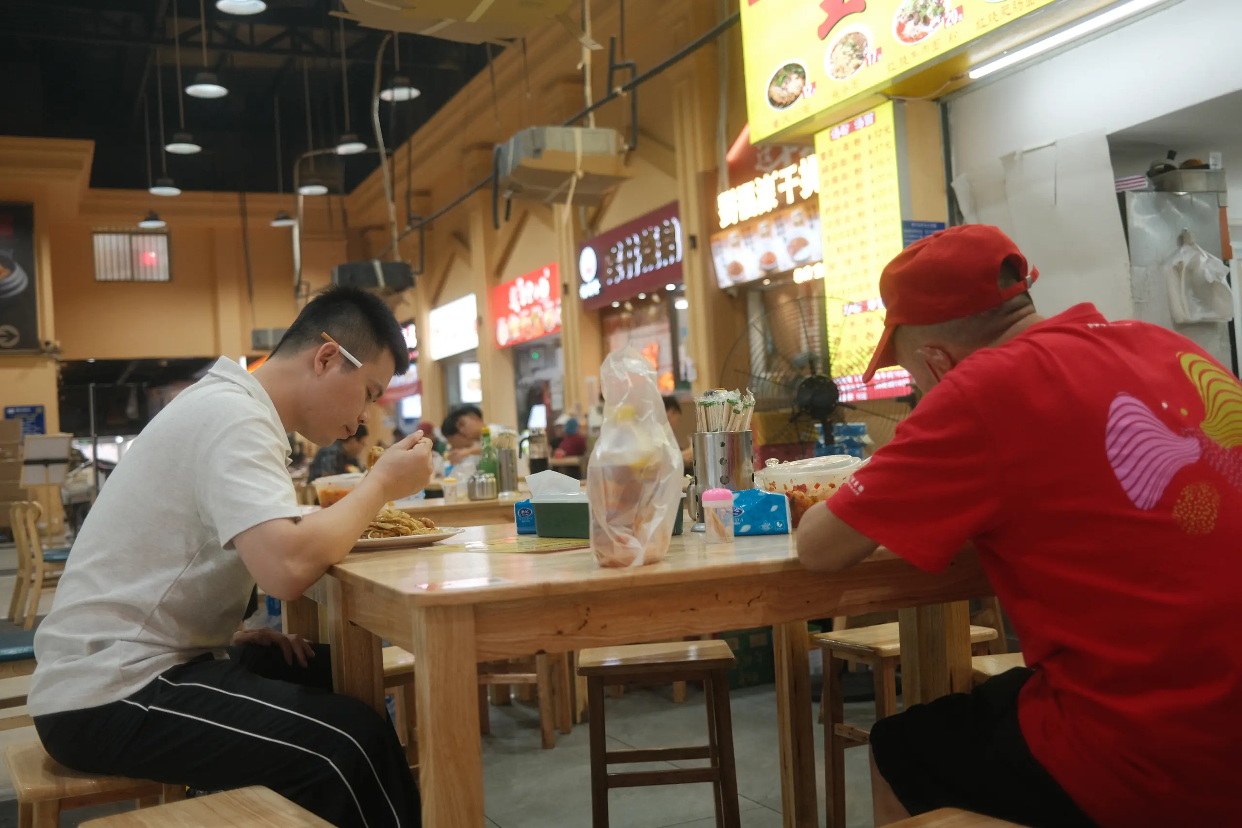 Two men eating at a wooden table in a food court