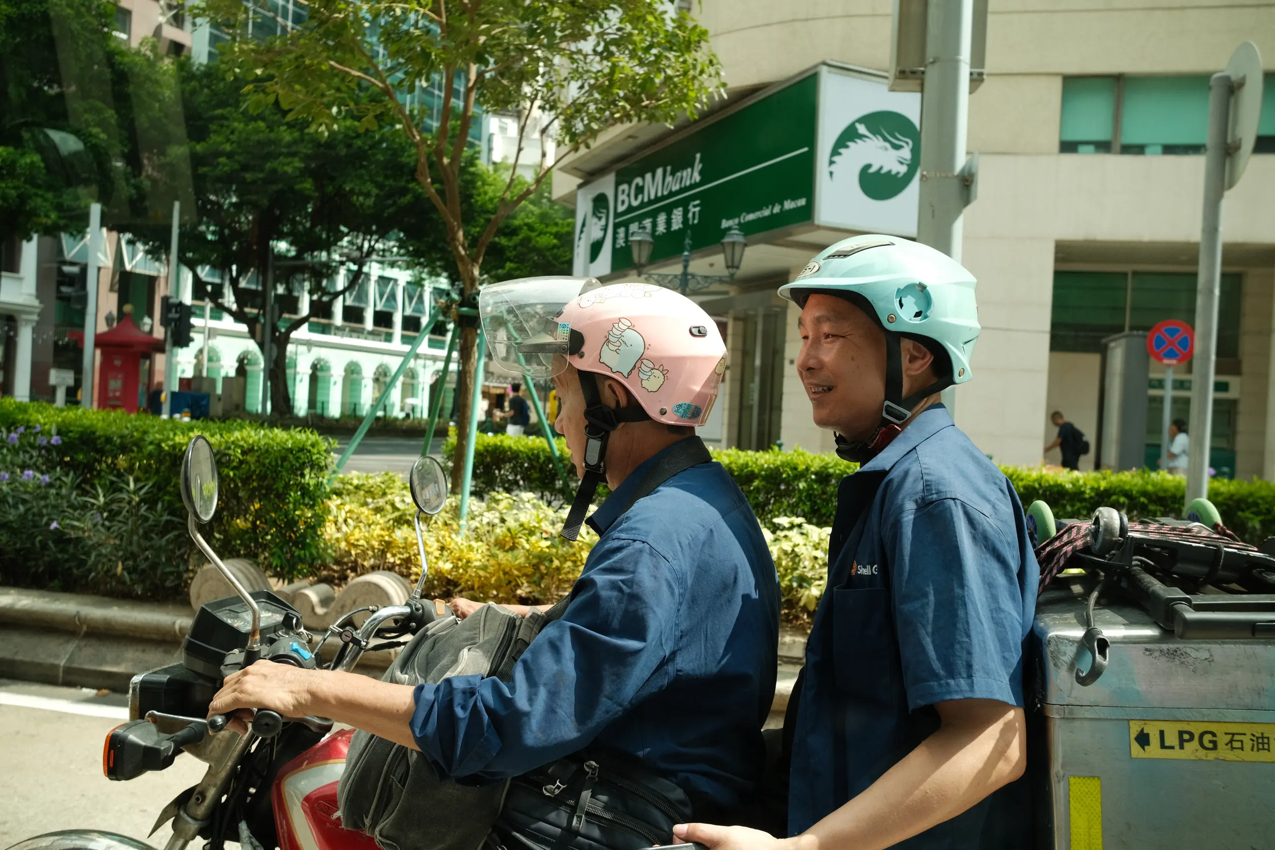 Two men in helmets riding a scooter past a hedge